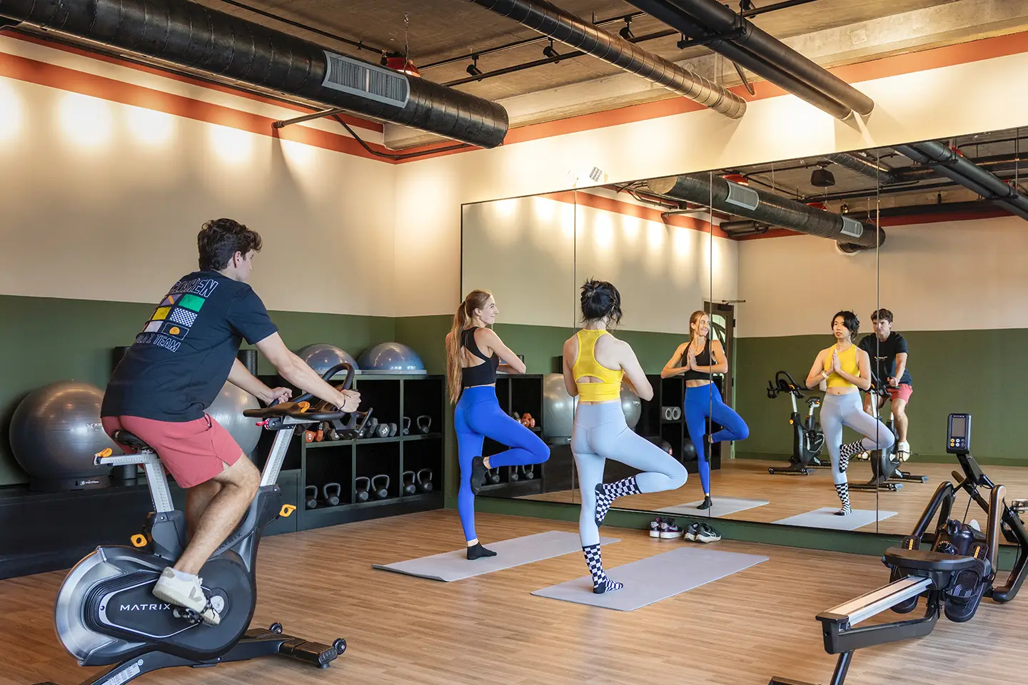 Fitness studio with two people doing yoga and another person on a stationary bike surrounded by weights and equipment.